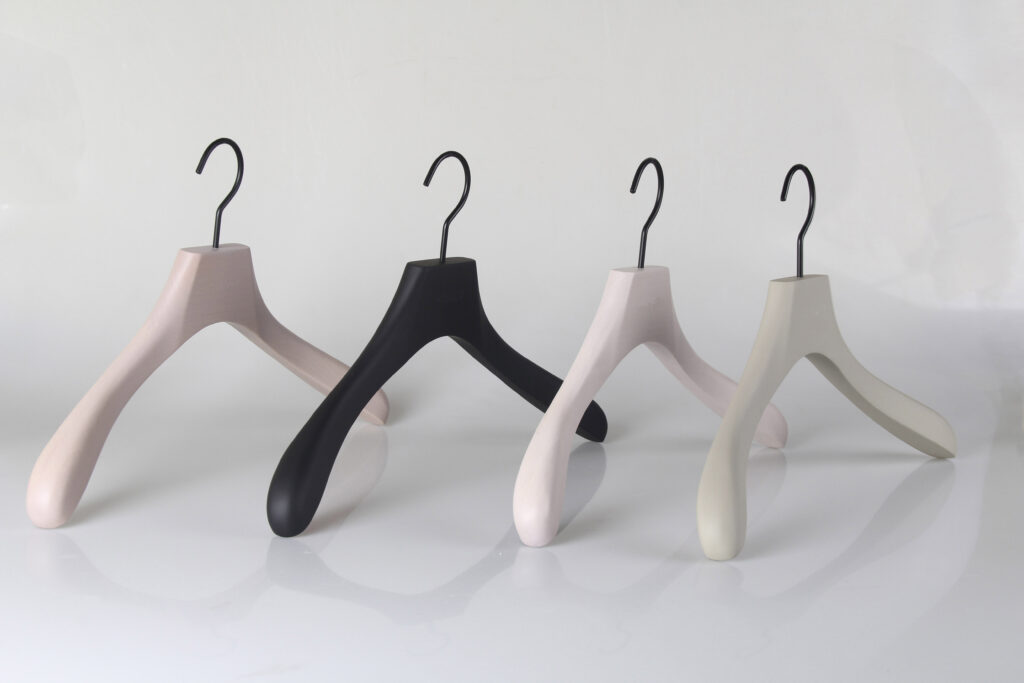 Hangers of London The image shows four modern and minimalist clothes hangers arranged in a row against a white background. The hangers are in neutral shades, including light pink, black, and beige, each featuring a thick, rounded design with metal hooks.