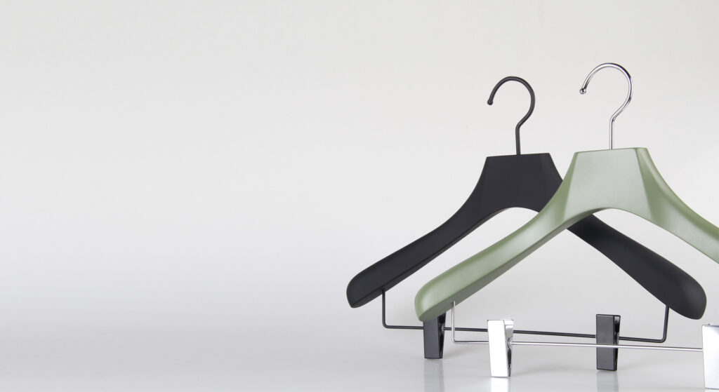 Hangers of London Two clothing hangers, one black and one green, are hanging on a white wall. The green hanger is placed in front of the black one, showing their simple and modern design. The background is minimal and clean.