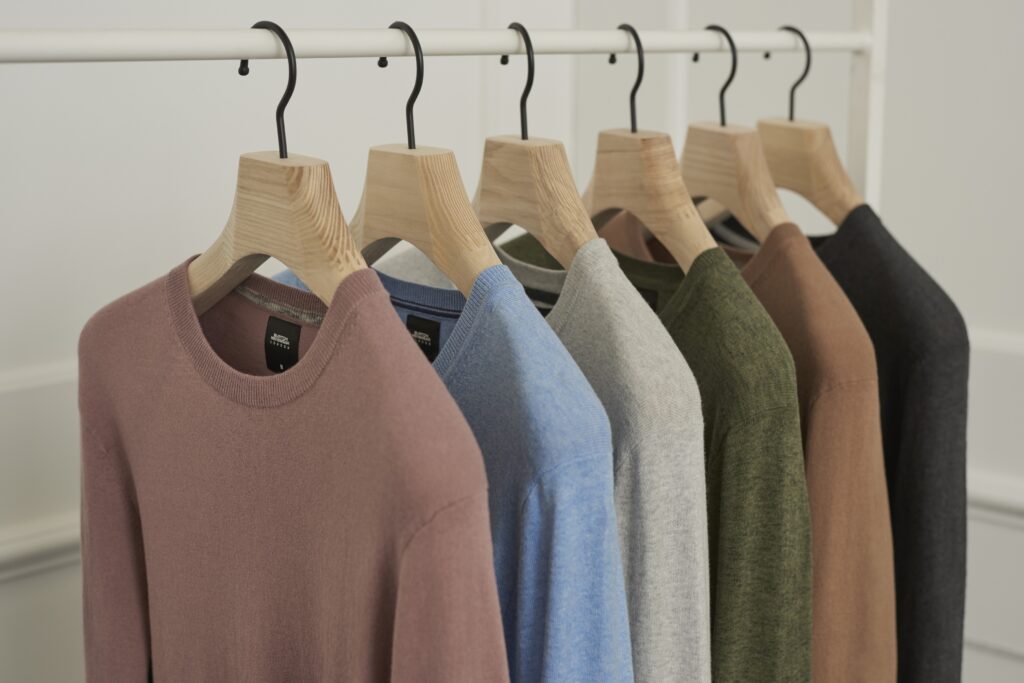 Hangers of London A row of six sweaters in various colors—pink, blue, gray, olive green, brown, and black—neatly hung on wooden hangers suspended from a white rack in a minimalist, white-walled room.